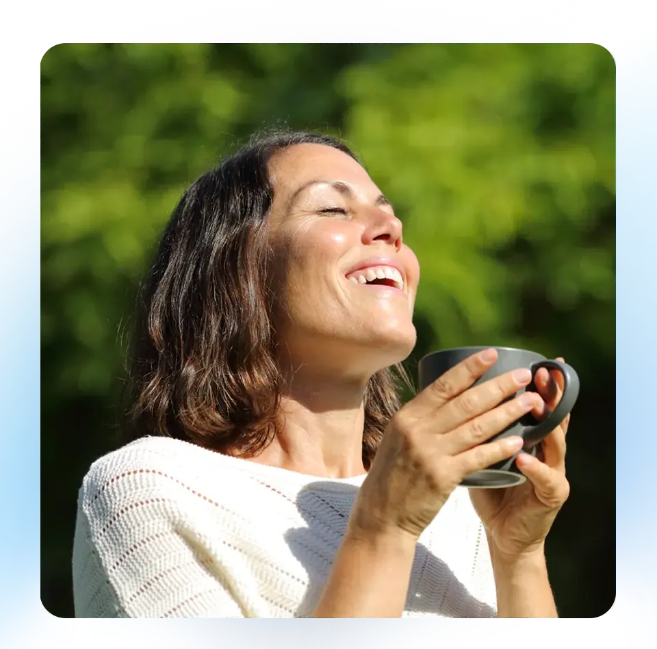 A woman smiling while she takes a deep breath and holds a coffee mug in her hands. Trees can be seen in the background. 