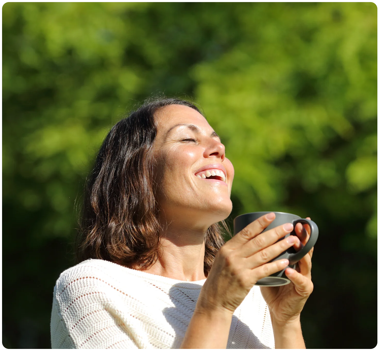 A woman smiling while she takes a deep breath and holds a coffee mug in her hands. Trees can be seen in the background. 