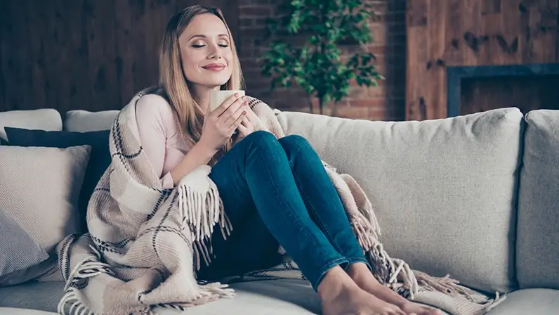 Kobieta siedząca wygodnie na kanapie, popijająca napój z kubka jako ukazanie objawów przeziębienia lub alergii.