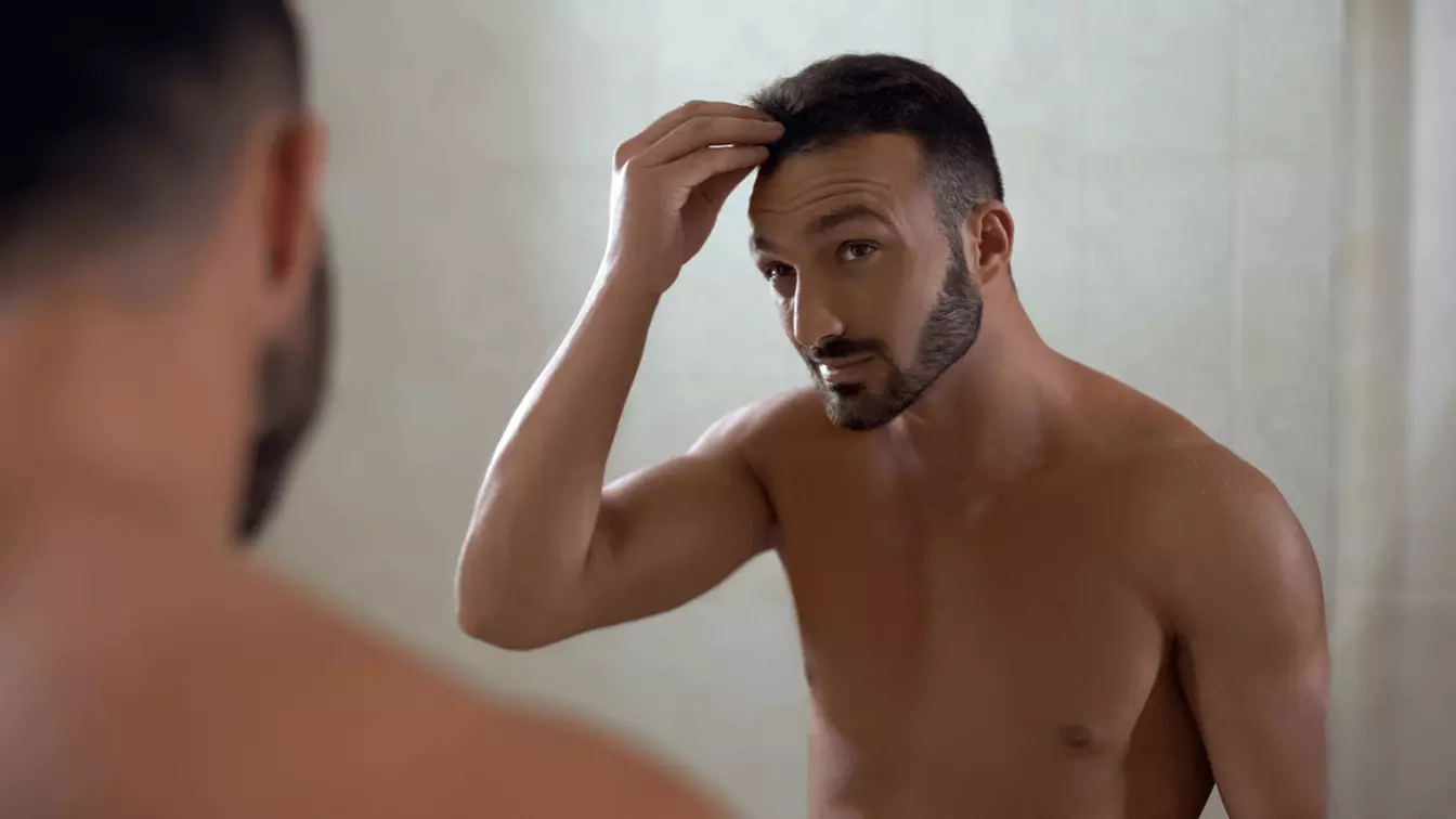 A man looking in the mirror, touching his hairline with his hand.
